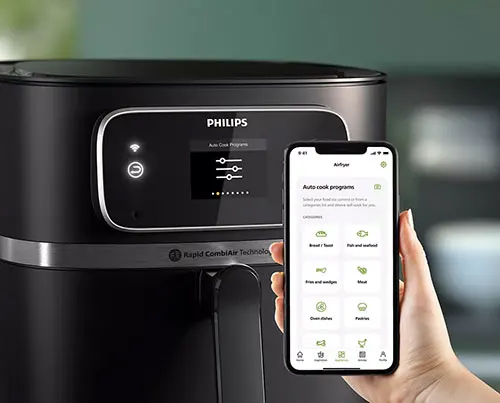 Friggitrice ad aria nera Philips con display digitale e mano che tiene un telefono con app di cucina.