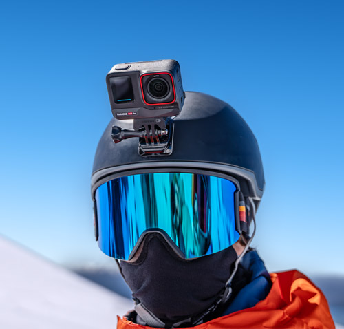 Persona con casco da sci nero e maschera blu, con action camera montata sopra. Cielo azzurro sullo sfondo.