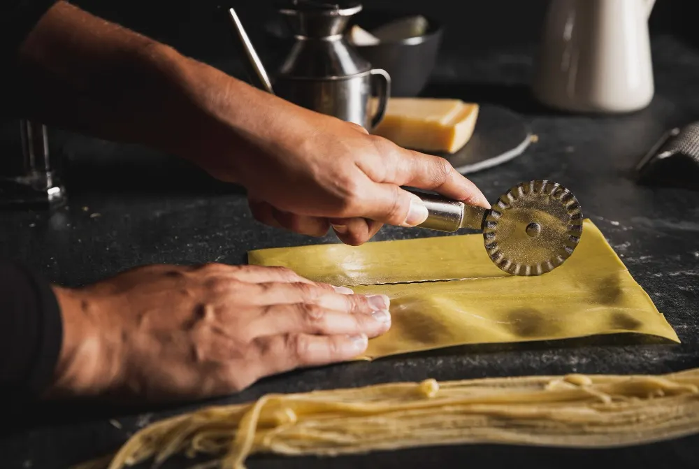Assortimento di pasta colorata su piatti e ciotole, con farina che forma la parola "PASTA" su un tavolo di legno chiaro.