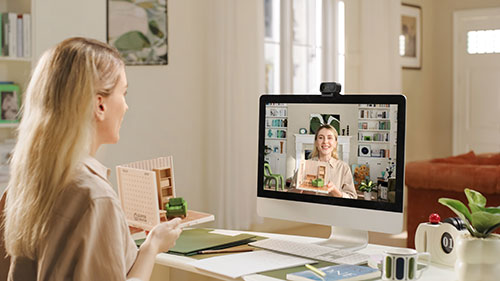 Donna che partecipa a una videochiamata su un computer desktop in un ambiente domestico. Sulla scrivania ci sono libri, piante e cancelleria.
