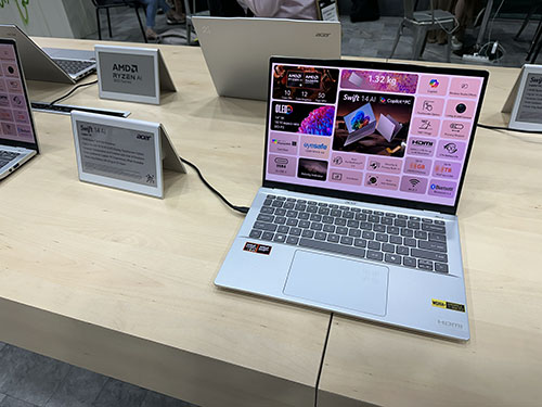 Un tavolo in legno chiaro espone laptop aperti e cartelli informativi. Schermi luminosi e tastiere grigie sono in primo piano.