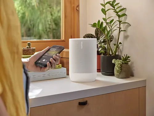 Vista di persona con telefono accanto a un altoparlante bianco su un mobile vicino a piante e finestra.