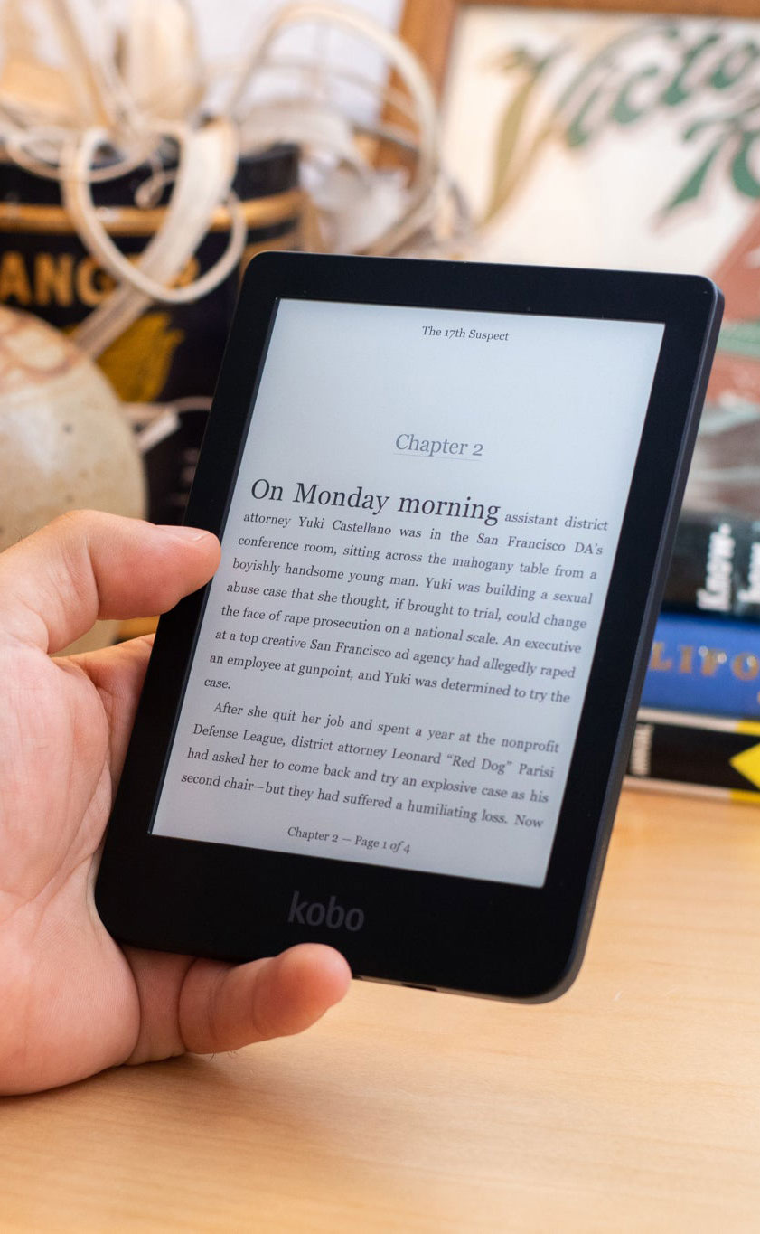 Mano che tiene un e-reader nero che mostra il capitolo 2 di un libro. Libri sfocati sullo sfondo.