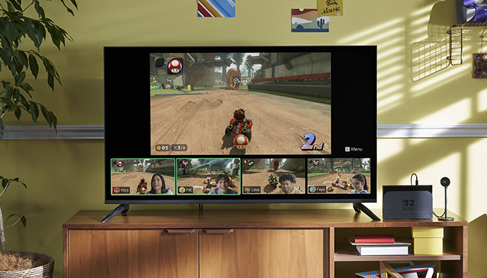 Una TV mostra Mario Kart con quattro giocatori visualizzati in basso. La TV è su un mobiletto di legno in una stanza con pareti gialle.