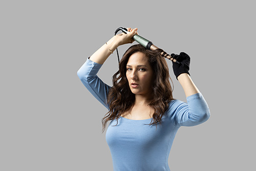 Donna con guanto nero arriccia i capelli con un ferro arricciacapelli. Indossa una maglietta blu. Lo sfondo � grigio.