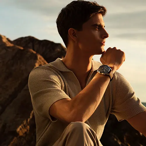 Uomo con maglia beige e pantaloni, indossa un orologio al polso, appoggiato alla mano; rocce sullo sfondo.