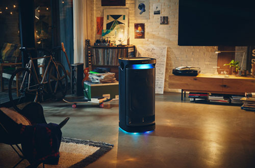 Un altoparlante cilindrico nero con luci blu illumina un soggiorno con TV, scaffali e dischi. Una sedia e un tappeto sono in primo piano.