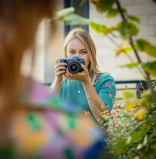 Una donna fotografa con una fotocamera nera, in un giardino con fiori e piante. Una figura sfocata � in primo piano.