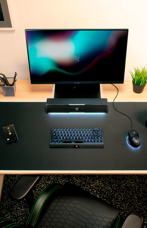 Scrivania con monitor, altoparlante, tastiera illuminata, mouse, cellulare, portapenne e pianta. Sedia da gaming in primo piano.