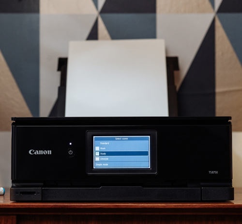 Una stampante nera Canon con uno schermo blu è su un tavolo di legno, con carta bianca inserita e uno sfondo geometrico.