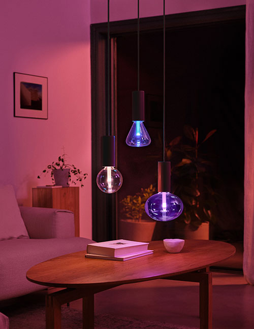 Interno illuminato con lampade a sospensione colorate sopra un tavolo ovale in legno con libri e ciotola. Divano e piante sullo sfondo.