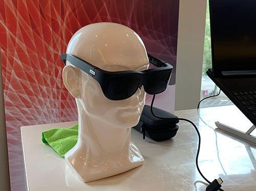 Manichino bianco con occhiali neri collegati a cavo. Sul tavolo, panno verde, custodia nera e laptop aperto. Sfondo rosso.