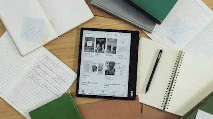 E-reader al centro, circondato da quaderni aperti, fogli con appunti, una penna e libri verdi su una superficie di legno.