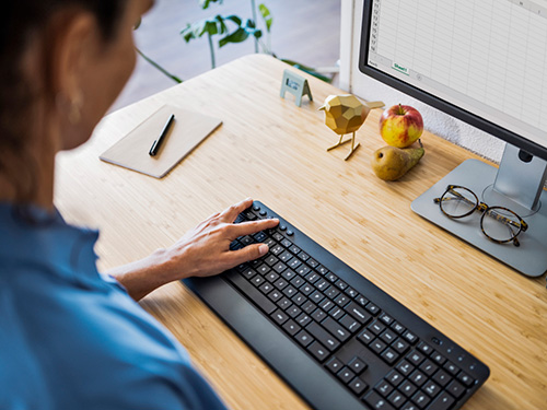 Donna che usa la tastiera del computer su una scrivania di legno con note, penna, frutta, occhiali e ornamento.