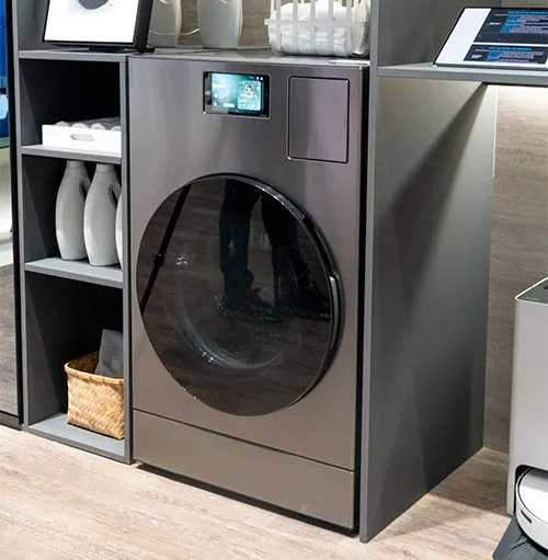 Lavatrice grigia con oblò nero e display digitale, affiancata da scaffali con detersivi e cesto di vimini.