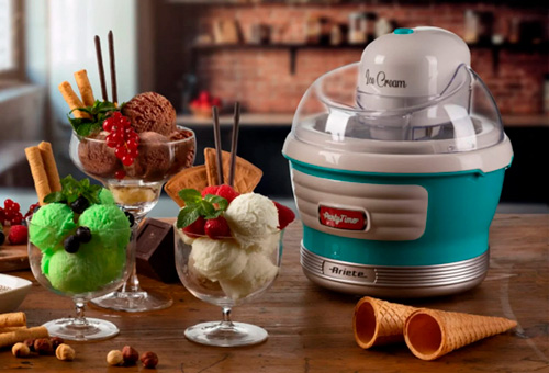 Gelatiera turchese accanto a tre coppe di gelato: verde, cioccolato e bianco, con coni e nocciole sul tavolo.