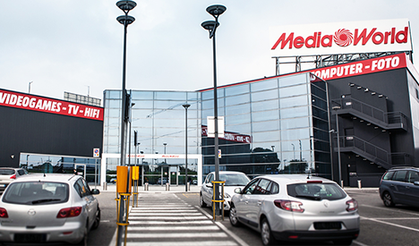 Esterno di MediaWorld con auto parcheggiate, segnaletica e insegne rosse su sfondo nero. L'edificio ha vetrate riflettenti.