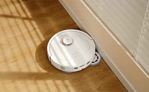 Aspirapolvere robot bianco vicino a una parete beige su un pavimento in legno marrone con ombre.