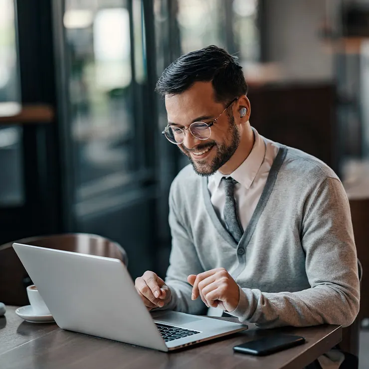 Uomo sorridente con occhiali lavora al laptop su un tavolo di legno. Ci sono una tazza bianca e un telefono nero accanto al computer.