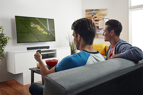 Due uomini seduti su un divano guardano una partita di calcio in TV. Uno tiene in mano un telecomando.