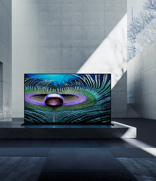 Un televisore mostra un'immagine astratta con viola, verde acqua e linee radiali su un supporto nero in una stanza illuminata.