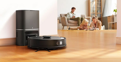 Aspirapolvere robot nero con base di ricarica su pavimento in legno. Persone sedute e uomo su poltrona sullo sfondo.