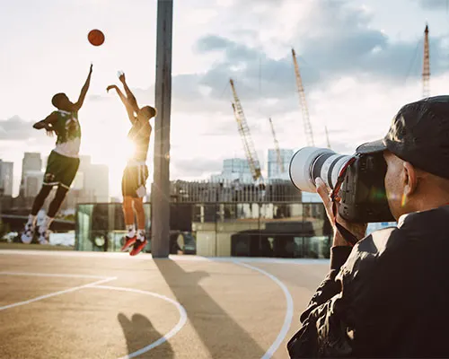 Fotografo con teleobiettivo riprende giocatori di basket su campo esterno con skyline urbano sullo sfondo.