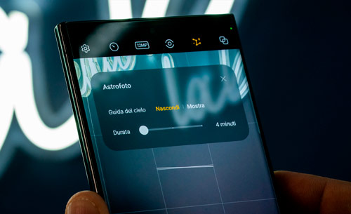 Primo piano di uno smartphone che mostra l'interfaccia di una fotocamera con impostazioni per l'astrofotografia. Uno sfondo con luci bianche.
