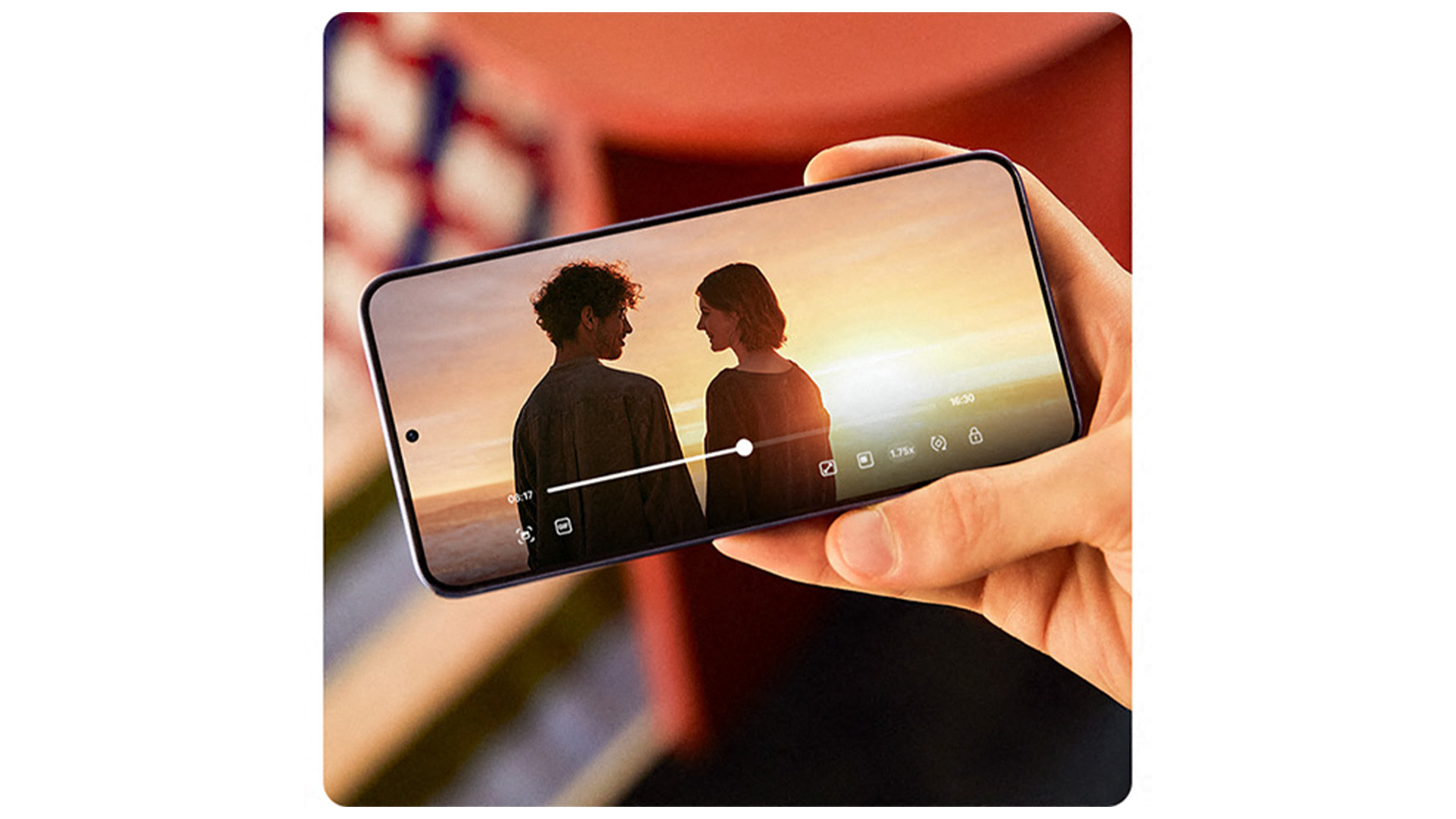 Una mano tiene un telefono che riproduce un video di due persone su uno sfondo arancione.