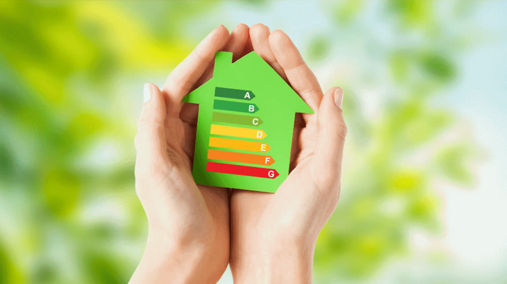 Due mani tengono un cartello a forma di casa verde con etichette colorate di efficienza energetica da A a G. Sfondo verde sfocato.