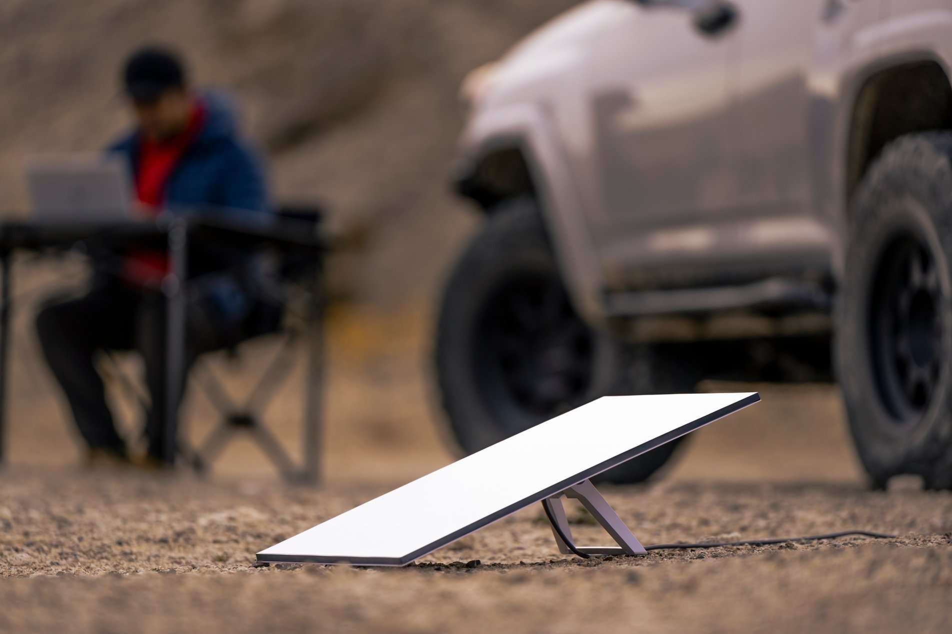 Antenna bianca su terreno sterrato, con auto e persona seduta a un tavolo sullo sfondo.