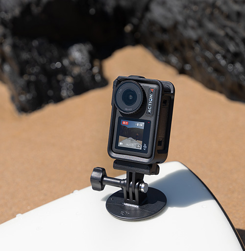 Una action camera nera su un supporto � posizionata su una superficie bianca con uno sfondo di sabbia e rocce scure.