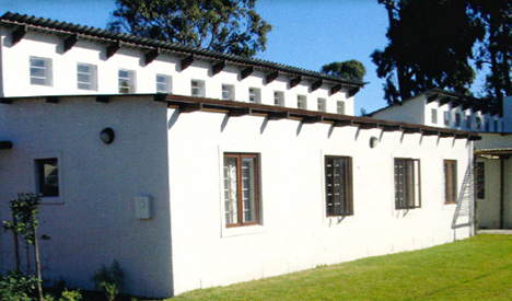 Edificio bianco a due piani con finestre rettangolari, tetto scuro e alberi sullo sfondo sotto un cielo azzurro.