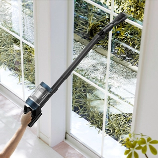Aspirapolvere nero pulisce una finestra bianca a riquadri. Si vedono piante verdi fuori dalla finestra e un muro bianco.