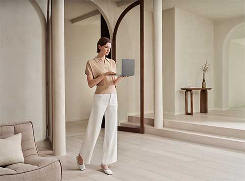 Donna in pantaloni bianchi che tiene in mano un laptop in una stanza con colonne e archi.
