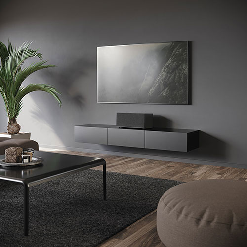 Soggiorno grigio con TV sopra mobile sospeso. Tavolino nero con oggetti decorativi, tappeto scuro e pouf chiaro. Pianta in vaso.