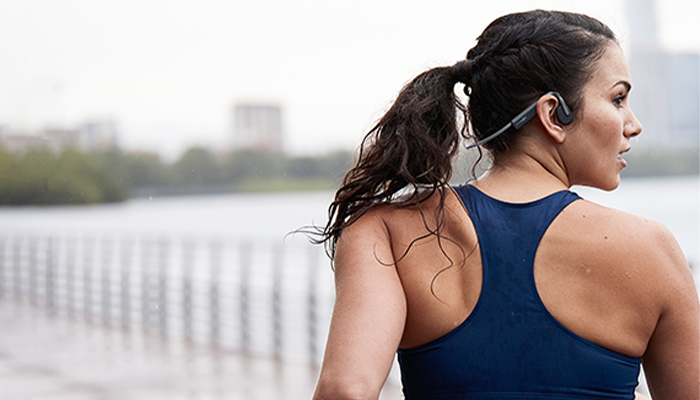 Donna con cuffie indossa canottiera blu, guarda a destra. Dietro, acqua, alberi e edifici sfocati.