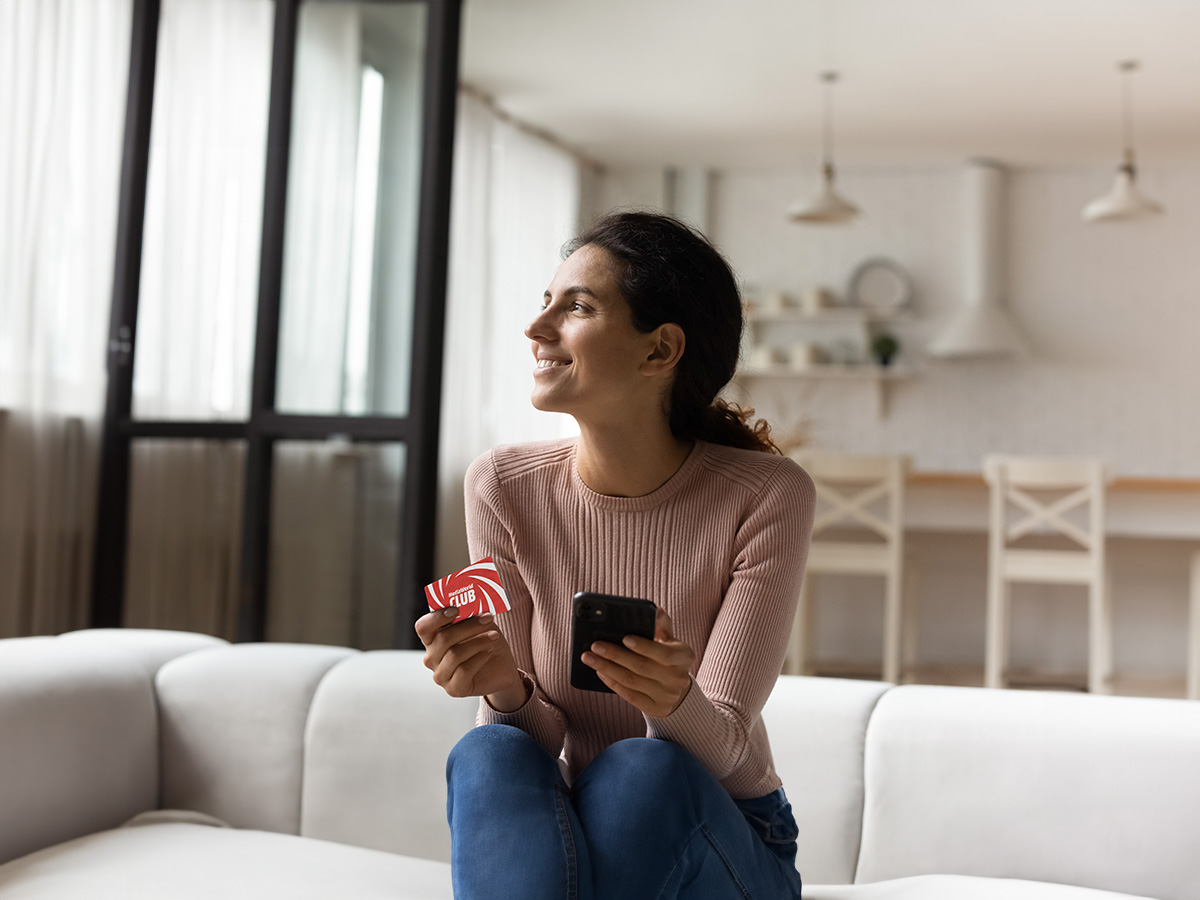 Donna sorridente seduta su un divano bianco con cellulare e carta club rossa. Sfondo con cucina e tavolo.