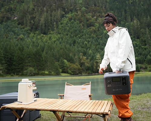 Donna in giacca bianca porta una power station vicino a tavolo di legno. Sul tavolo ci sono macchina da caff� e sedia. Alberi e lago sullo sfondo.
