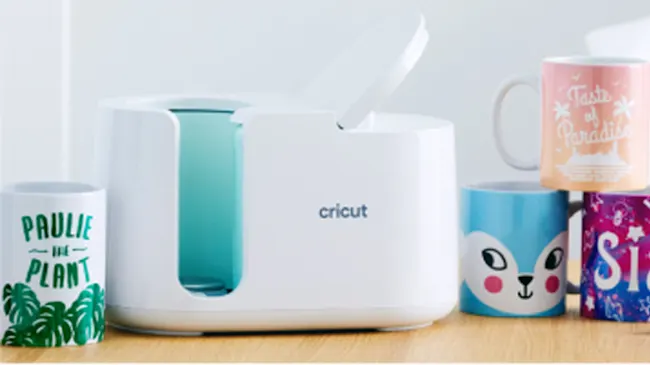Macchina Cricut bianca con tazze decorate su un tavolo di legno chiaro e sfondo bianco.