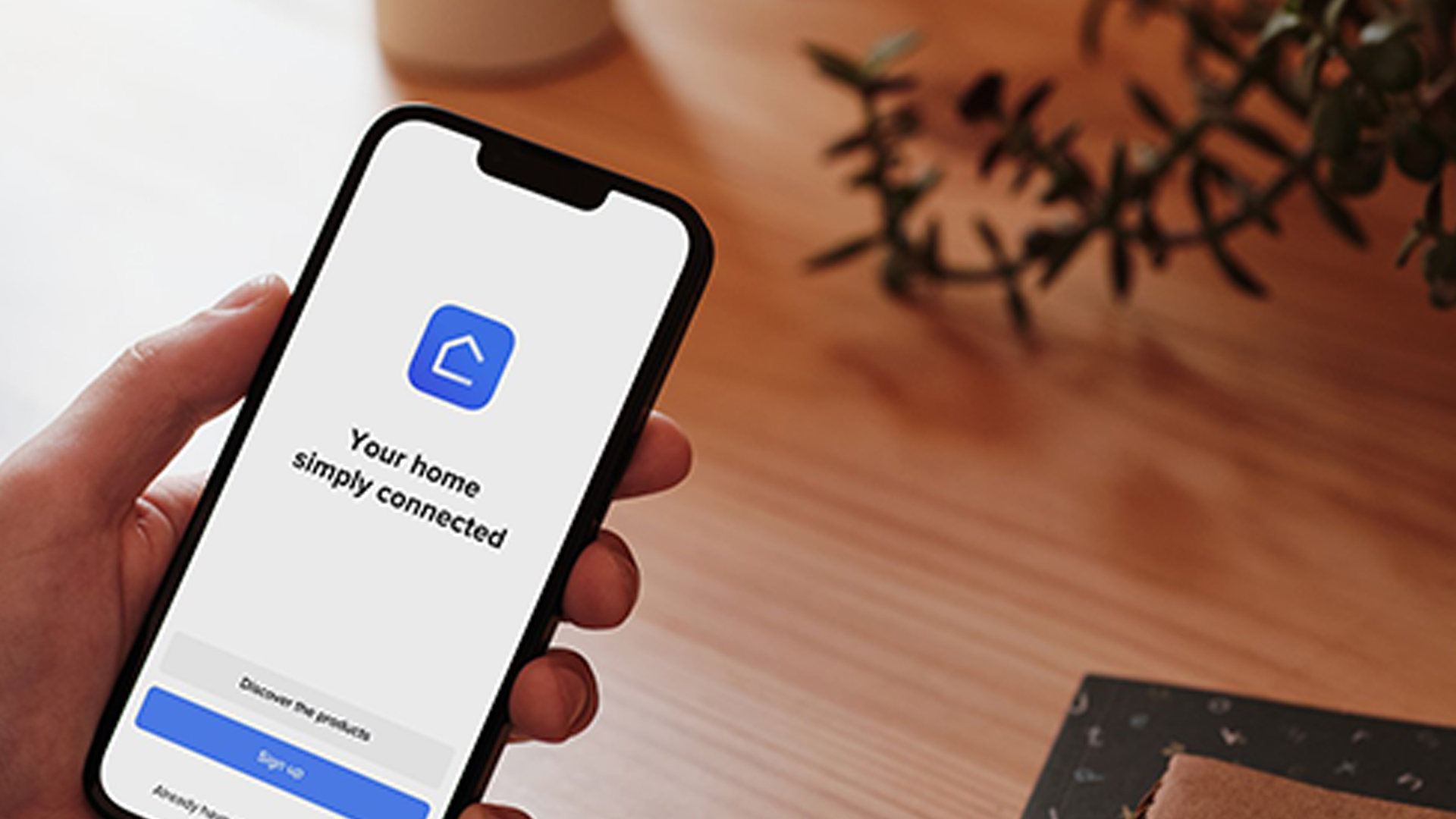 Mano che tiene un telefono con app smart home. Scritta: "La tua casa semplicemente connessa". Sullo sfondo, pianta e tavolo di legno.