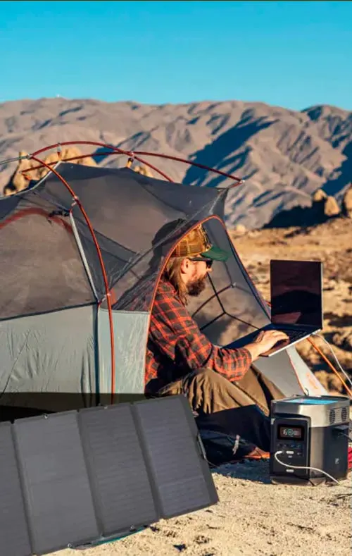 Uomo in tenda grigia usa un laptop. Un pannello solare nero e una power station sono visibili su un terreno desertico con montagne.