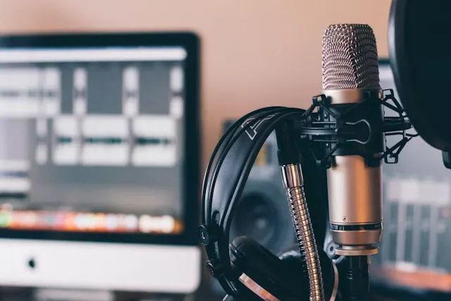 Microfono dorato su supporto, cuffie nere, cavo argentato e schermo di computer con software audio in secondo piano.