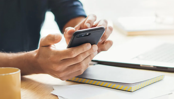 Uomo che tiene un telefono nero sopra un taccuino su una scrivania con una tazza e un laptop.