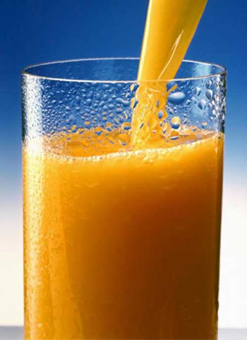 Versando liquido arancione in un bicchiere trasparente con gocce d'acqua su sfondo blu.