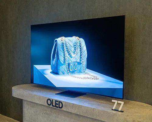 TV OLED su tavolo beige con borsa blu. Sul tavolo, la scritta "OLED" e il numero "77". Sfondo scuro.