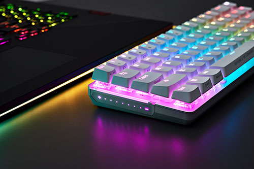 Tastiera bianca retroilluminata con luci colorate accanto a un laptop nero su una superficie scura.
