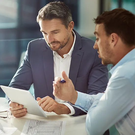 Due uomini discutono documenti su un tavolo. Uno in giacca blu tiene un foglio, l'altro gesticola con una penna in mano.
