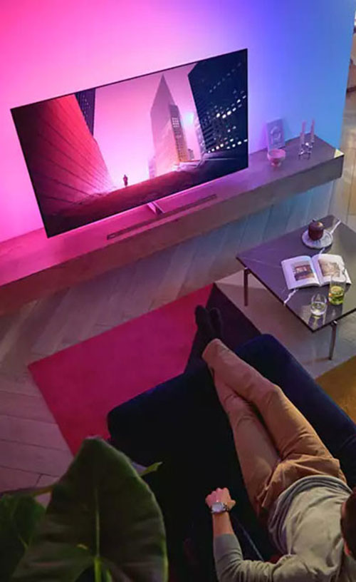 Vista dall'alto di persona rilassata su divano blu, TV con skyline illuminato, tavolino con libro aperto.