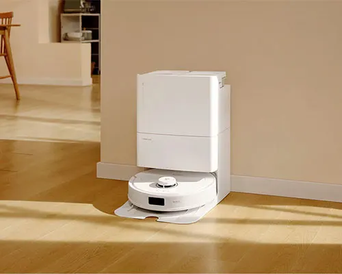 Aspirapolvere robot bianco con stazione di ricarica su pavimento di legno chiaro, vicino al muro beige.